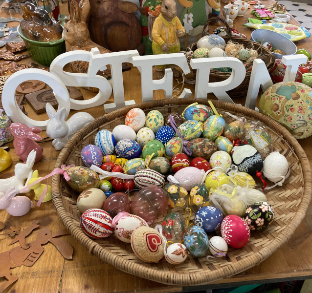 Ein flacher geflochtener Korb voller bunter Hühner-Glaseier, dahinter in weissen Lettern der Schriftzug "Ostern" auf dem Ostertisch im Brocki, alles Material aus Haus- und Wohnungsräumungen